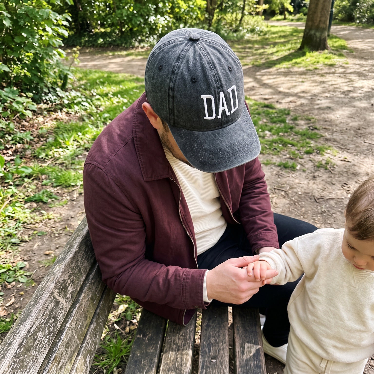Dad in a Black DAD cap seated on a park bench holding his child’s hand