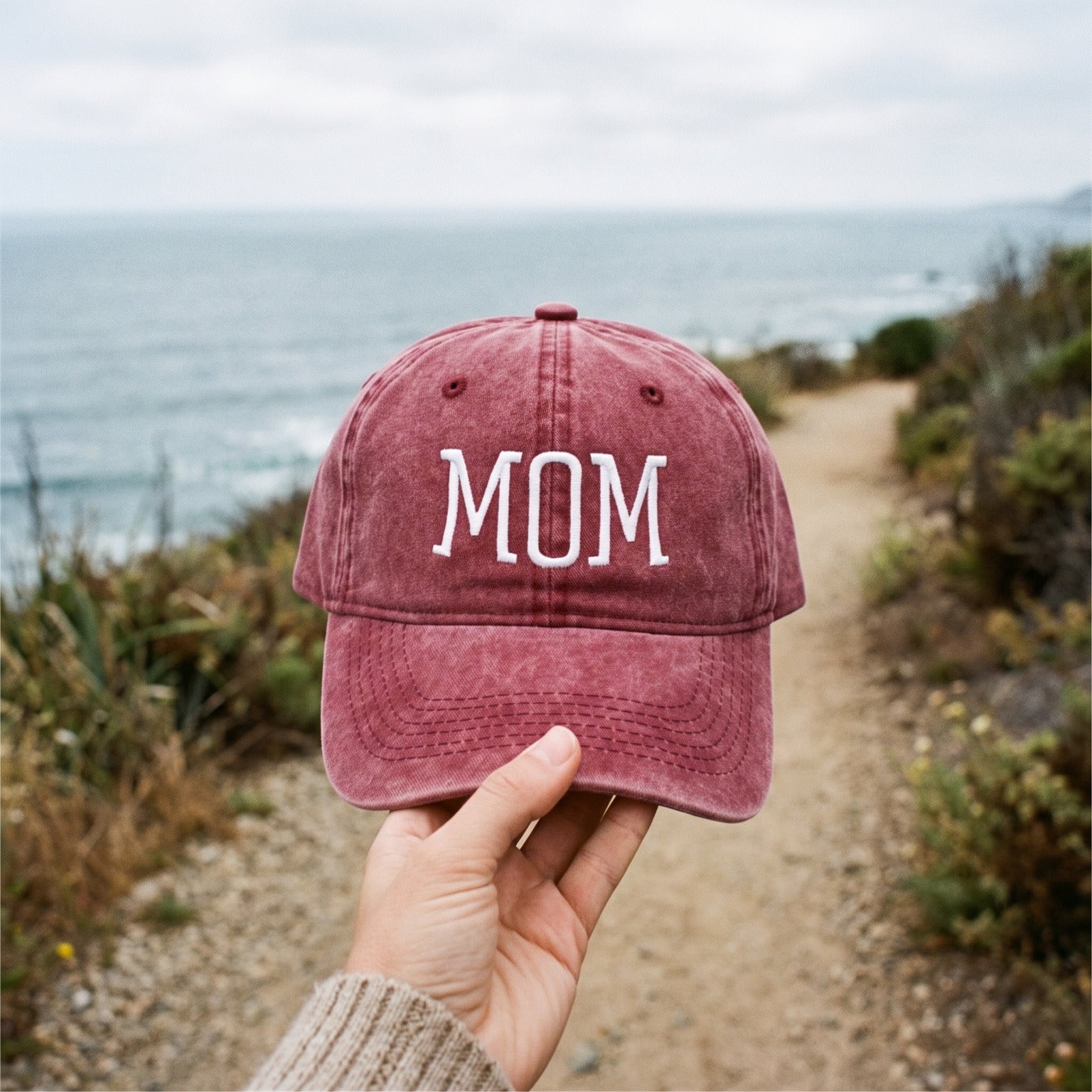 Hand on the brim of a Red MOM cap by the coast